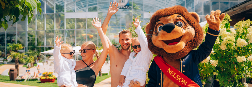 Eltern mit zwei Kindern stehen gemeinsam mit Nelson im Thermengarten und jubeln.