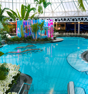 Large thermal pool with turquoise blue water, green palm trees and a colorfully lit sauna can be seen in the background.