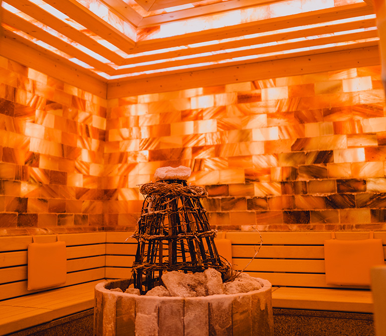 Interior view of a stylishly lit sauna with orange glowing salt walls and a sauna heater in the center.