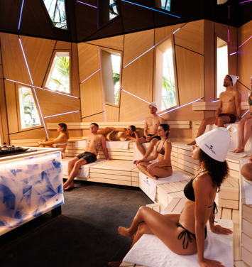 Several guests in swimwear sit on wooden benches in a sauna surrounded by neon lights.
