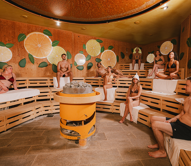 People in a sauna decorated with lemons and leaves on the walls; one person is performing an Aufguss in the center