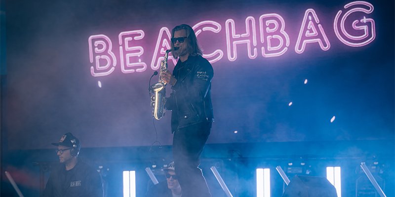 Musiker mit Sonnenbrille spielt Saxophon auf Bühne vor leuchtendem Schriftzug 'BEACHBAG'