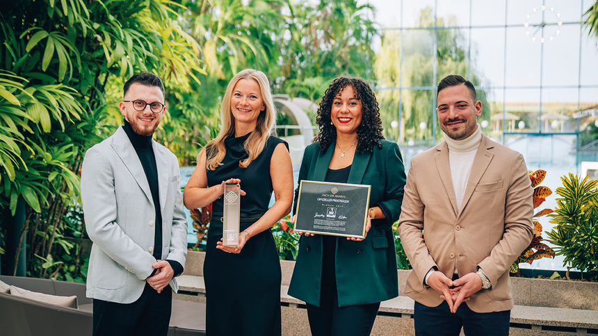 Vier Personen stehen vor einer Glaswand mit Pflanzen, zwei Frauen halten einen Award und eine Urkunde, alle lächeln