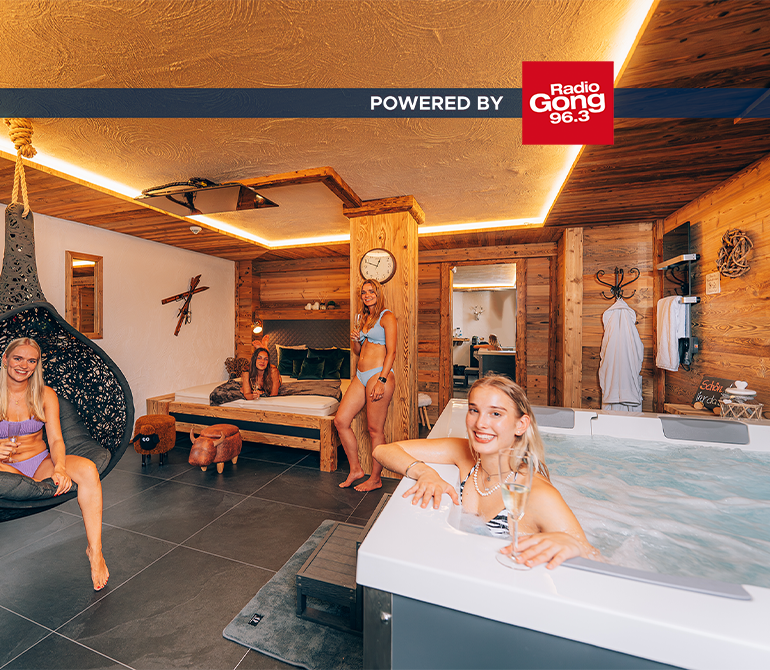 Eine Frau im Whirlpool mit Sektglas, im Hintergrund stehen zwei weitere Freundinnen am Bett. Der Raum ist gemütlich im Holz-Hüttenstil eingerichtet.