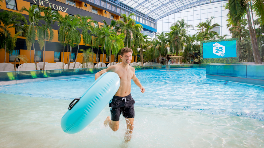 Thomas Müller läuft mit blauem Schwimmreifen im Wellenbad mit Palmen und geöffnetem Glasdach