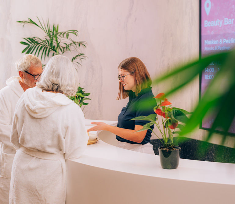 [Translate to en:] Eine Mitarbeiterin der Therme gibt Gesichtsmasken an zwei Gäste aus.