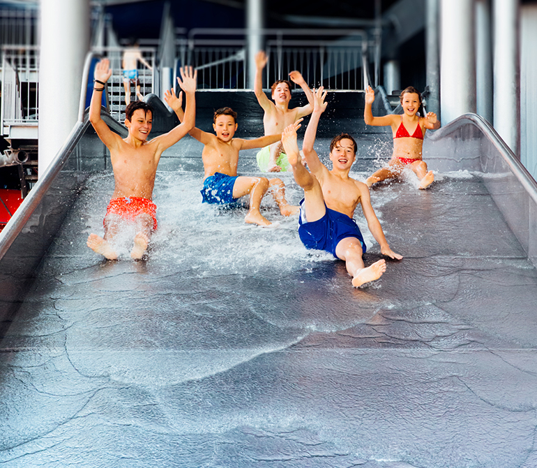 Five children slide down a wide water slide side by side, laughing and waving.