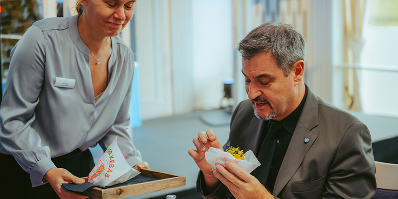 Mann in dunklem Anzug isst einen Döner, während eine Frau mit hellblauer Bluse und Namensschild ihm ein Tablett mit weiteren Dönern hält