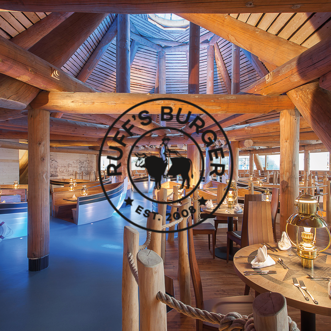 The interior of a restaurant featuring rustic wooden beams, round tables with lamps and benches, a blue floor, and skylights