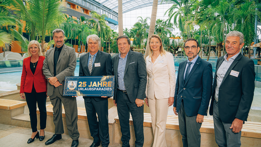 Sieben Personen in formeller Kleidung stehen in einer Halle mit Palmen und einem Pool. Zwei Personen halten ein Schild mit der Aufschrift '25 Jahre Urlaubsparadies'.