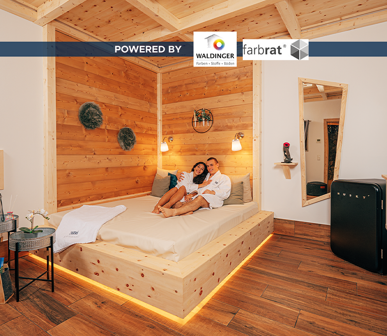 Ein Paar entspannt auf einem großen Bett mit vielen Kissen und Decke in einem gemütlich beleuchteten Raum im Holz-Hüttenstil.