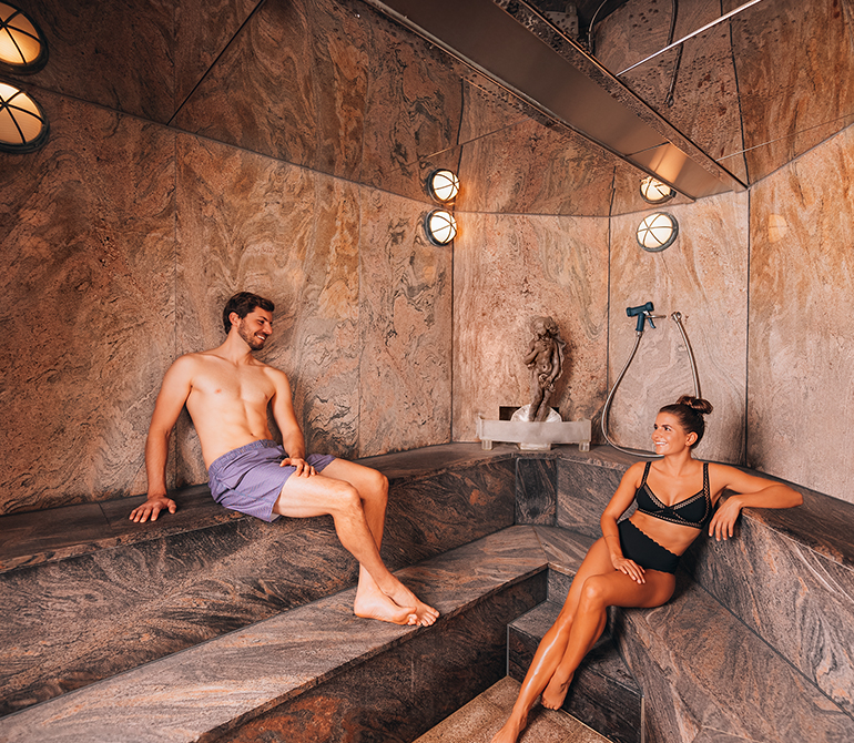 A man and a woman relax in a Roman steam room with marble walls and benches, decorated with a statue and round wall sconces