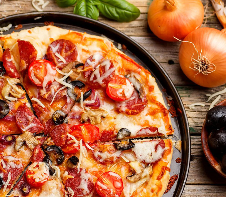 Pizza with salami, cherry tomatoes, olives, and cheese on a black plate, surrounded by onions and basil on a wooden table
