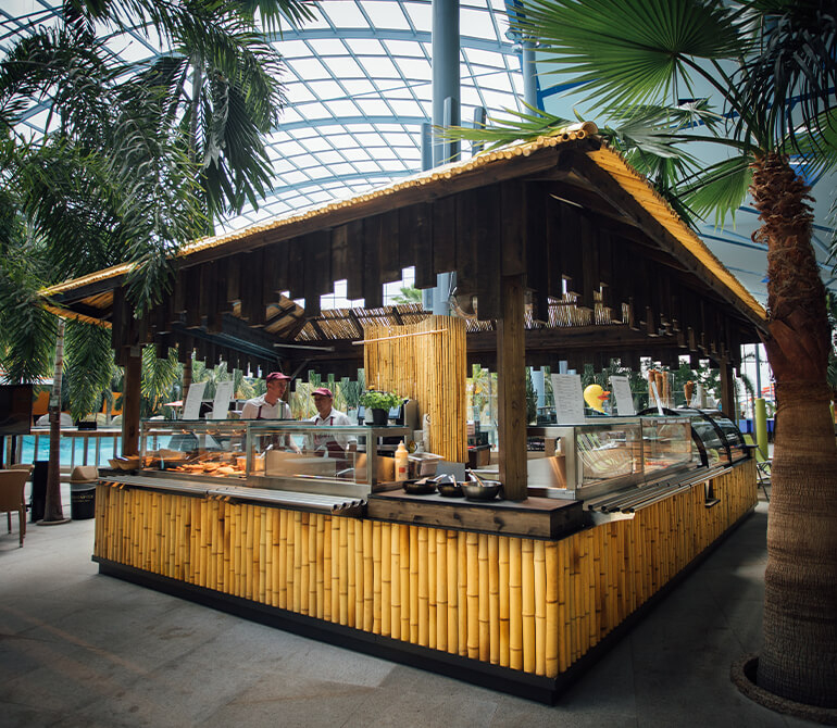 A bamboo-clad food stand with a thatched roof in a tropical setting featuring palm trees and a glass roof at the Nelsons Beach Restaurant at Therme Erding