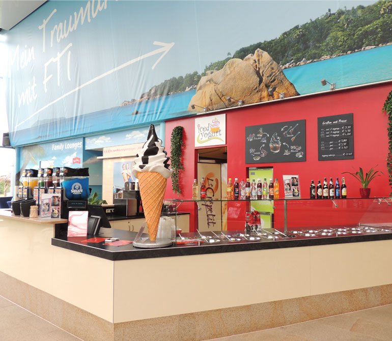 Counter at an ice cream or frozen yogurt shop featuring a large ice cream cone display, various bottles and toppings, a red wall with menu boards, and a beach scene in the background