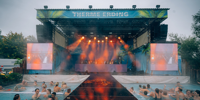 Außenbühne mit Schriftzug 'Therme Erding' und beleuchtetem Laufsteg, davor Menschen im Thermalbecken bei Veranstaltung