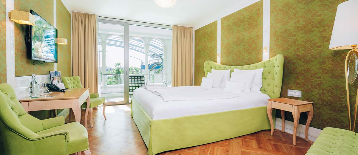 Helles Hotelzimmer mit grünem Interieur, großem Bett, Schreibtisch, Sesseln und Zugang zu einem Balkon mit Blick nach draußen