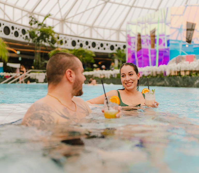 Two people holding drinks with straws at the bar, surrounded by tropical plants. Closes daily at 10:30 p.m.