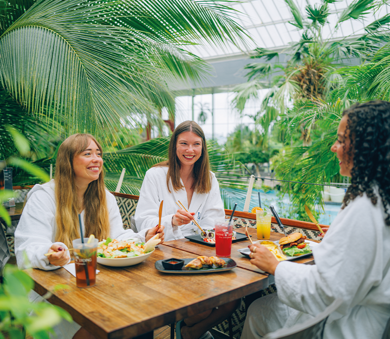 Three women in white bathrobes are clinking glasses, sitting at a wooden table with food, surrounded by green plants