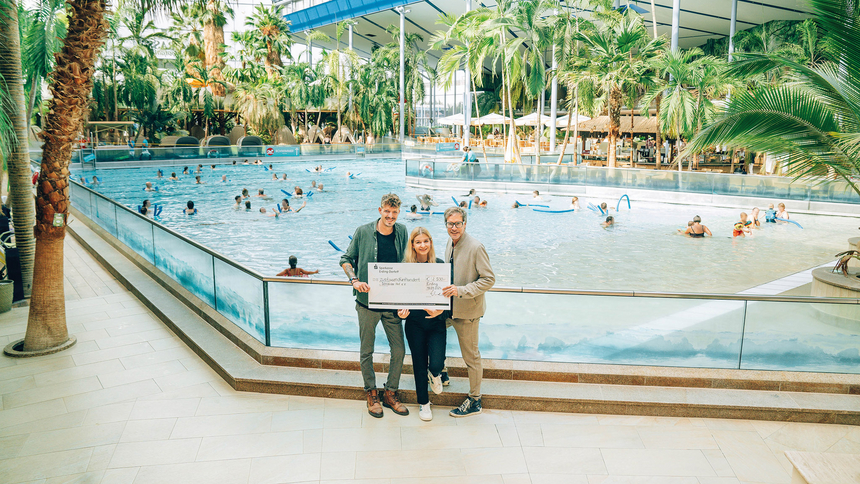Drei Personen stehen in einem Hallenbad und halten einen großen Scheck, im Hintergrund schwimmen Menschen im Wasser