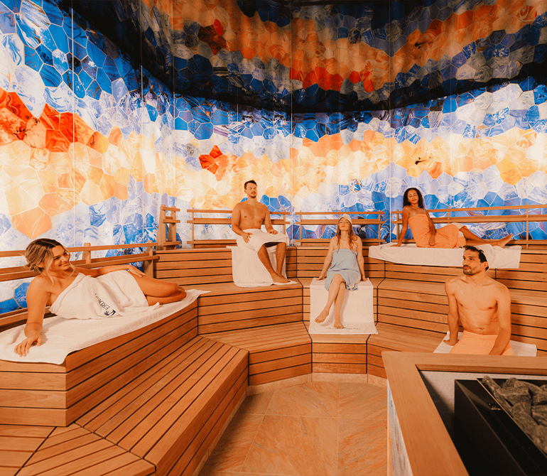 Mehrere Gäste entspannen in einer modernen Sauna mit farbenfrohen Mosaikwänden und warmem Licht.